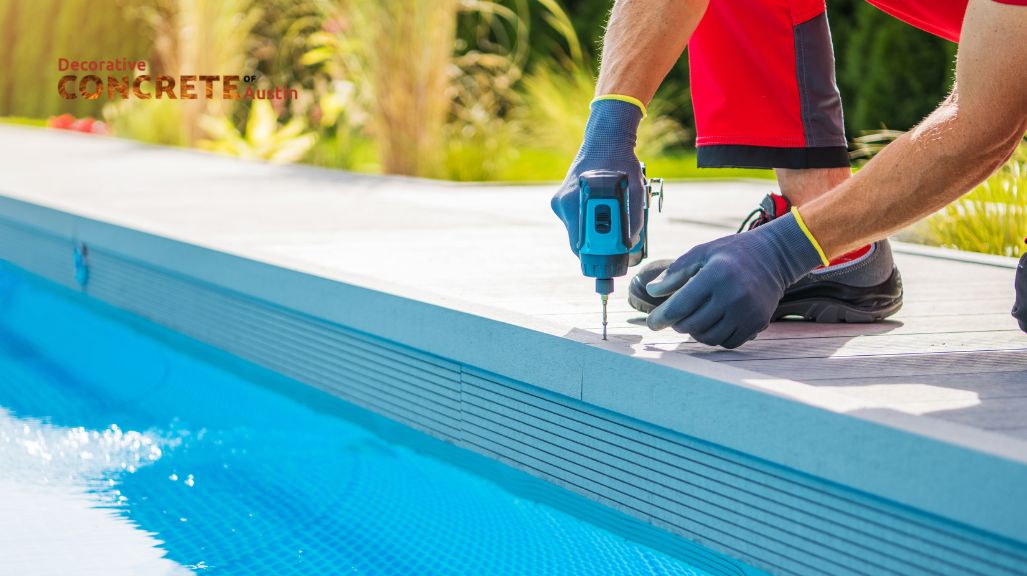 A person in red shorts and gloves uses a power drill on a pool deck