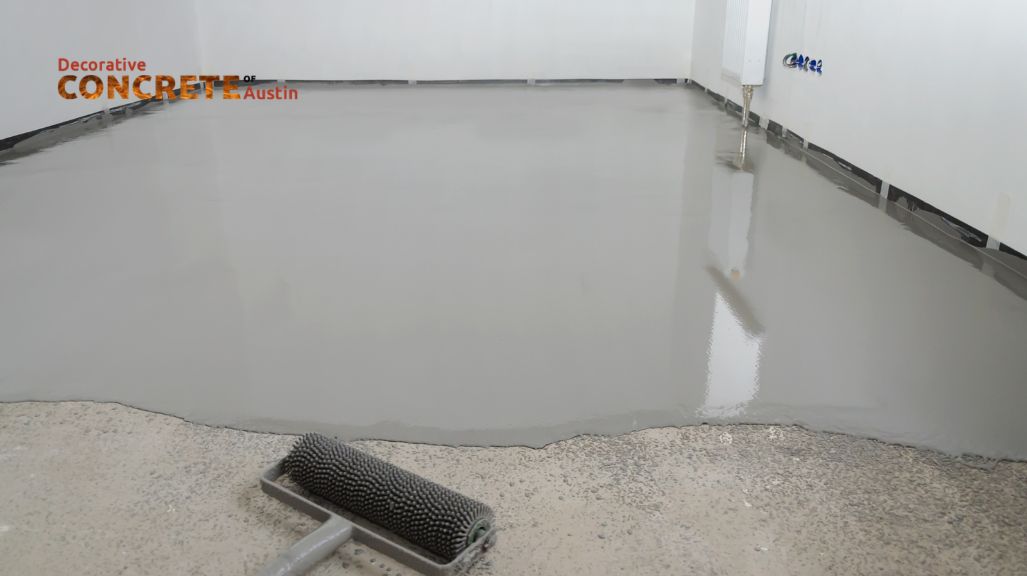 A worker in overalls and a red shirt kneels on a concrete floor, smoothing it