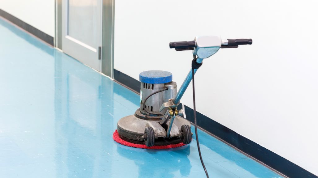 A floor buffer with a red pad is placed on a shiny blue floor, near a glass door.