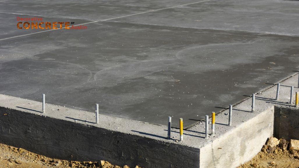 A smooth, gray concrete foundation slab with protruding metal rebar is set on a sandy, unfinished construction site, conveying a sense of progress.