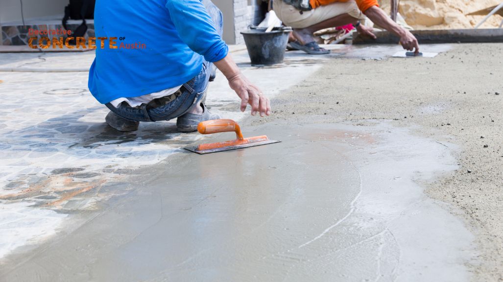 A worker in a blue shirt kneels while smoothing wet concrete with a trowel.