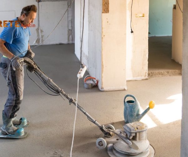 A worker in a blue shirt smooths concrete with a floor sander in a partially renovated room.